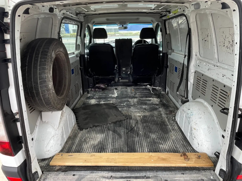 Van interior with a tire, wooden board, and scattered items on a dirty floor.