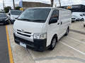 White Toyota van parked in a parking lot with other vehicles and buildings in the background.