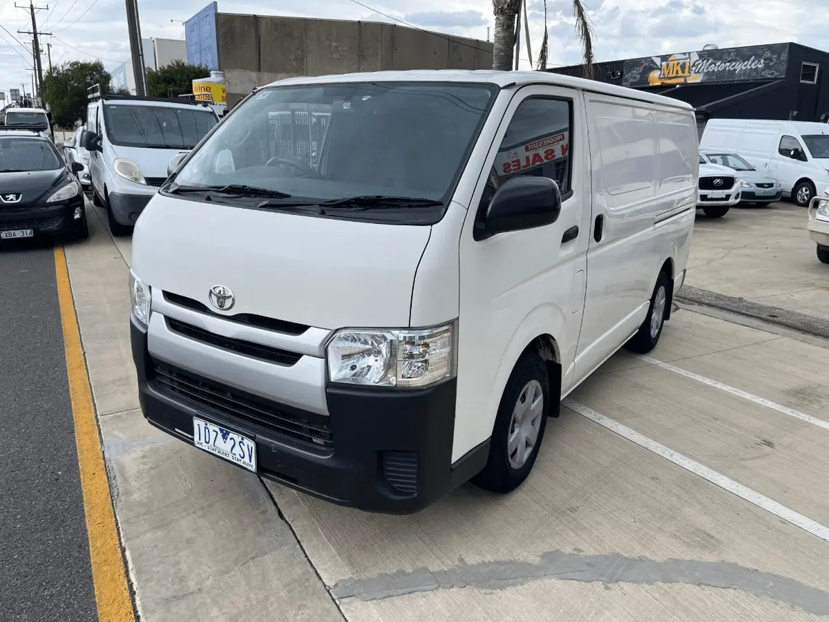 White Toyota van parked in a parking lot with other vehicles and buildings in the background.